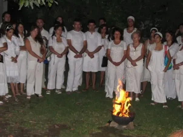 De noche en el jardín, un grupo numeroso de personas vestidas de blanco forma un semicírculo de pie; en primer plano arde el fuego en un brasero de hierro apoyado sobre la tierra.