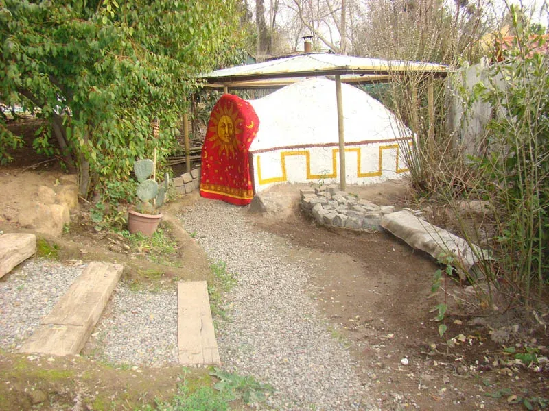 La cúpula de barro del temazcal, encalada en blanco, con una greca escalonada pintada en amarillo recorriendo su base y un textil rojo con un sol dorado cubriendo la entrada. Alrededor hay tierra, gravilla y árboles.