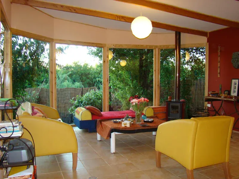 Un espacio interior vidriado y abierto al jardín, con sillones amarillos y una mesa baja de madera con comida servida.
